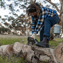 Person in a plaid shirt and jeans wearing black pull-on work boots, using a chainsaw to cut a log in an outdoor rural setting. Person in a plaid shirt and jeans wearing black pull-on work boots, using a chainsaw to cut a log in an outdoor rural setting.