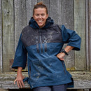 Smiling person in a blue, slightly muddy outdoor jacket leans against a rustic wooden wall. The scene conveys a casual, outdoorsy, and relaxed vibe.