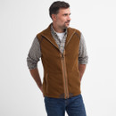 Man poses in brown fleece gilet and plaid shirt, hands in pockets, looking to the side. Casual, relaxed style set against a plain background.