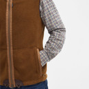 A person wearing a brown gilet over a plaid shirt is shown from the shoulders to waist, hands in pockets, conveying a casual and cosy style.