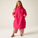 Girl smiling in a vibrant pink long coat with furry cuffs, paired with checkered pants and flip-flops. She stands confidently on a plain backdrop.