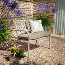 Garden bench with a cushion surrounded by purple flowers and a potted plant in sunlight.