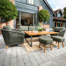 A modern garden lounge set with green-cushioned seating, two footstools and a wooden coffee table, arranged on a patio outside a house with open bi-fold doors and topiary trees.