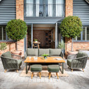 A contemporary outdoor lounge set arranged on a paved patio, featuring a three-seater sofa, two armchairs, two footstools and a wooden coffee table, styled in front of open bi-fold doors and neatly shaped topiary trees.