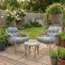 Two grey cushioned wicker chairs with a glass-top table sit on a patio surrounded by potted plants and lush greenery, evoking a serene garden retreat.