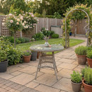 A serene garden features a round glass table with a vase of flowers, surrounded by potted plants on a stone patio. Roses bloom near a wooden fence.