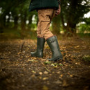 A child walks through a forest wearing green boots and rolled-up brown pants. The ground is covered with leaves and twigs, creating a rustic autumn vibe.