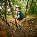 Young boy with red hair, wearing a plaid shirt, green vest, and rubber boots, explores a lush forest, looking up with curiosity.
