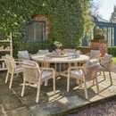 An outdoor dining scene featuring a round wooden garden table with eight rope-weave armchairs, arranged on a stone patio beside a brick house covered in green foliage.