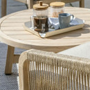 A close-up of a round wooden outdoor coffee table holding a tray with coffee, mugs and biscuits beside a rope-weave armchair.