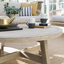 A close-up of a round wooden outdoor coffee table styled with mugs, a teapot and a plant, with a cushioned rope-weave sofa in the background.