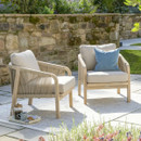 Two rope-weave outdoor armchairs with soft beige cushions positioned on a stone patio beside a rustic garden wall.