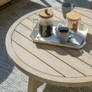 A wooden slatted round outdoor coffee table in sunlight, styled with a tray containing a cafetière, mugs and biscuits on a textured outdoor rug.