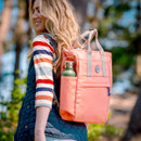 A person carrying a coral tote bag with a water bottle pocket, dressed in a striped shirt, outdoors.