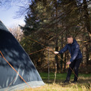 A person in a blue jacket secures a tent with orange cords in a forest clearing on a sunny day, evoking a sense of adventure and calm.