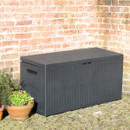 Closed black rattan-effect garden cushion storage box on a cobbled patio next to a brick wall and potted plant.