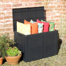 Black garden storage box with the lid open, filled with colourful outdoor cushions beside a brick wall.