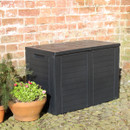 Closed black garden cushion storage box placed on a cobbled patio next to a brick wall and potted plants.
