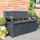 Black garden storage bench with slatted back and armrests on a patio, positioned against a red brick wall with potted plants.