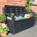 Black garden storage bench with the lid open, revealing garden tools and cushions stored inside, set on a paved patio beside a brick wall.