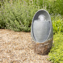A textured stone water feature in an oval shape sits on gravel. It has a smooth, egg-like sphere atop, surrounded by lush green foliage in a serene garden setting.
