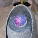 A glowing orb emitting purple and pink light sits atop a textured, stone-like fountain. A potted plant is blurred in the background, suggesting a serene setting.