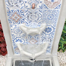 Ornate mosaic-tiled fountain with blue and white patterns. Water flows gently into three tiers, surrounded by green leaves and white pebbles, creating a serene ambiance.