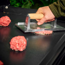 Hand using a smash burger press to flatten mince patties on a hot griddle.