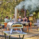 Ooni outdoor pizza oven with chimney producing smoke while people gather in a garden dining setting in the background.