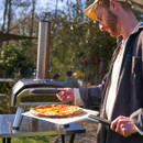 Person placing a freshly cooked pizza on a peel in an Ooni outdoor wood-fired pizza oven in a garden setting.