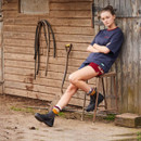 Person in a t-shirt and shorts sitting on a chair by a wooden barn with a pitchfork nearby.