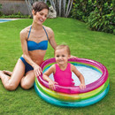 Mother sitting on grass beside a toddler playing in a small inflatable rainbow paddling pool in a garden.