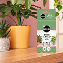 A package of Miracle-Gro Drip & Feed for potted plants on a wooden table, surrounded by a green plant in an orange pot and a small succulent.