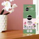 Box of Miracle-Gro Drip & Feed plant food on a wooden table next to a white orchid in a textured vase. The scene conveys a fresh, nurturing tone.