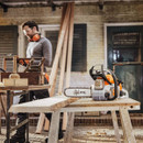 A man wearing ear protection works with wood in a workshop. A chainsaw is on a table beside him. The scene is industrious and focused.