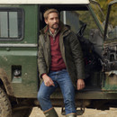 A man in a green jacket and jeans leans casually against an off-road vehicle in a rugged, outdoors setting. He wears a thoughtful expression.