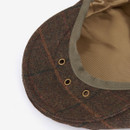 Close-up image of a brown tweed flat cap's interior, showing its plaid pattern and grommet details. The fabric lining is a contrasting beige.