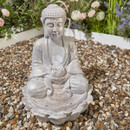 Stone-effect seated Buddha water feature holding a lotus bowl, shown on decorative gravel with flowers behind.