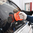 A gloved hand uses an orange suction lifter to lift a car window in a garage setting. The reflection of tools and shelves is visible in the glass.