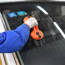 A person in blue overalls uses an orange suction tool on a car windscreen. The atmosphere is focused, suggesting repair or installation work.