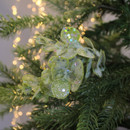 Close-up of leaves covered in glitter and sequins, nestled among branches of a Christmas tree with soft, warm white lights blurred in the background.