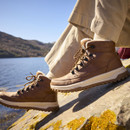 Brown fleece-lined hiking boots worn on rocks beside a mountain lake, featuring lace-up fastening and chunky outdoor soles.