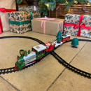 Close-up of a Christmas train set with festive locomotive and Santa carriage travelling on black track beneath a decorated Christmas tree.