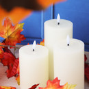 Close-up of three white LED pillar candles with flame-shaped lights, displayed with red and orange autumn foliage.