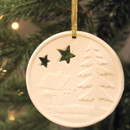 A ceramic white disc with a wintery scene of a lodge, tree and cut out stars imprinted onto it on a grey background with a christmas tree and lights