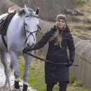 A person in a dark grey coat leading a white horse with a bridle.