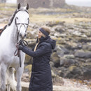 Person in a dark grey coat and matching headband holding reins of a white horse against a rocky landscape.