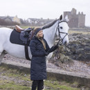 A person in a dark grey coat stands next to a white horse with a castle in the background.