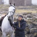 Person in dark grey coat and matching headband holding reins of a white horse, with a rocky background.