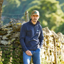 Man outdoors wearing a navy Narvik fleece, leaning against a stone wall in a countryside setting.